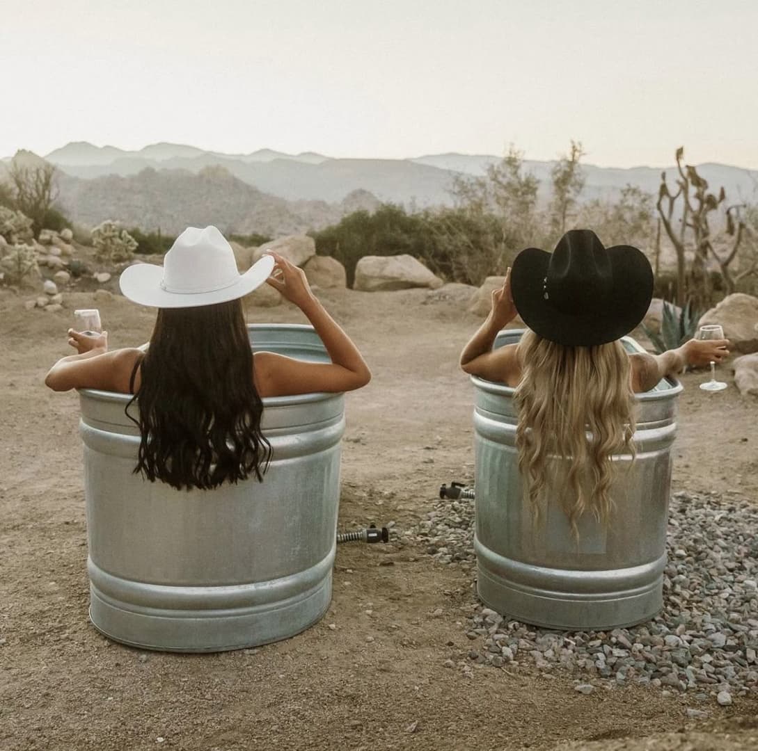 Two women in cowboy hats relaxing in metal stock-tank tubs in the desert